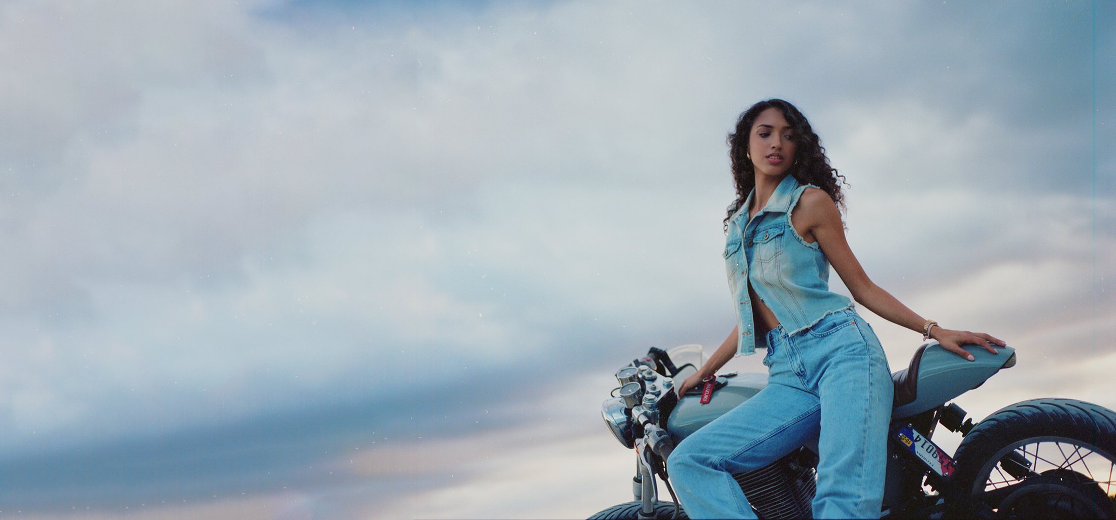 Model on a motorcycle against a wide overcast sky.